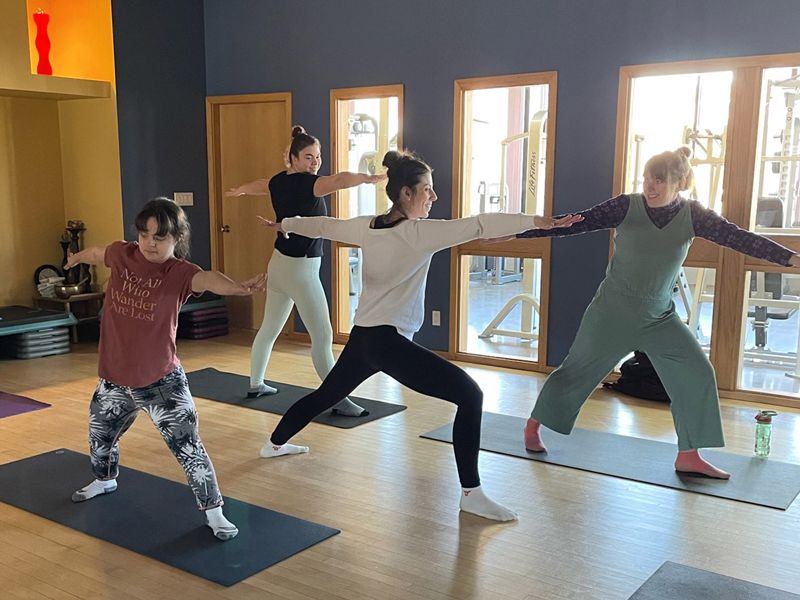 Adaptive-Yoga_1 Four people practise yoga together on mats in a bright studio, arms extended in a warrior pose, creating a calm, supportive space for wellness and connection.