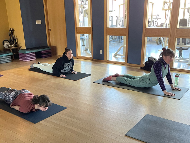 Adaptive-Yoga_2 Three participants practise gentle yoga on mats in a bright studio, holding cobra/upward-dog and resting poses in a calm, supportive wellness session.