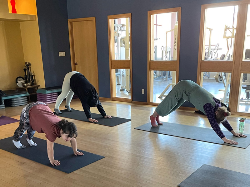 Adaptive-Yoga_4 Three participants practise yoga in a bright studio, holding downward-dog poses on mats, reflecting a calm, supportive HopeBridges-style wellness session.