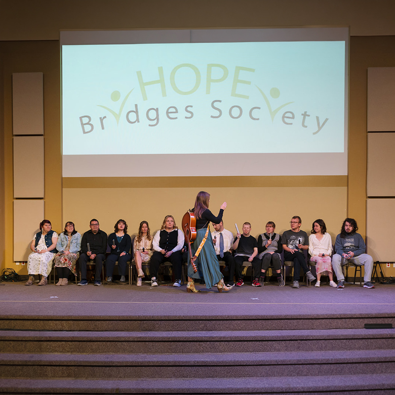 Wide shot of a stage in an auditorium. A woman holding an acoustic guitar stands centre‑stage facing a seated group of people arranged in a row of chairs behind her. Above them, a large projected screen displays the ‘HOPE Bridges Society’ logo. Microphone stands and cables are visible at the front of the stage, with steps leading down to the audience area.