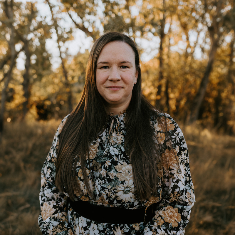 Portrait of Brettyn-Rose, a contributor to Hope Bridges community concerts. She is a woman with long, straight brown hair, wearing a dark dress with large autumnal floral patterns and a wide black belt. She smiles gently at the camera, standing outdoors in a field with blurred trees and warm, golden sunlight in the background.