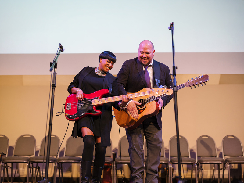 Hope Bridges – Artists Among us Gala Two musicians stand on stage holding a red electric bass and an acoustic guitar, with microphone stands and chairs behind them and a Hope Bridges Society banner projected above.