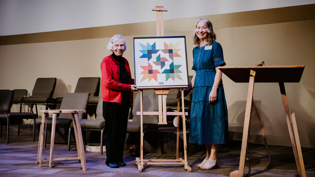 Hope Bridges – Artists Among us Gala Two people stand on a stage beside an easel displaying a framed colourful geometric artwork during HopeBridges (hopebridges.ca) past honours recognizing Christene Howard.