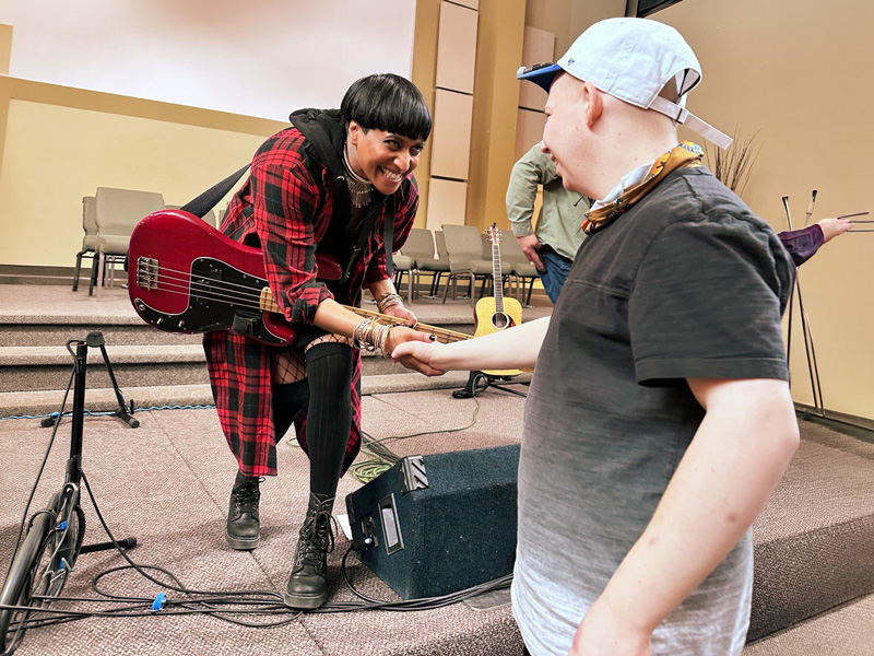 Community-Concert-Music-Series-1 A smiling musician holding a red bass guitar leans forward from a carpeted stage to warmly shake hands with a young person wearing a backwards light blue baseball cap, sharing a joyful moment of connection.