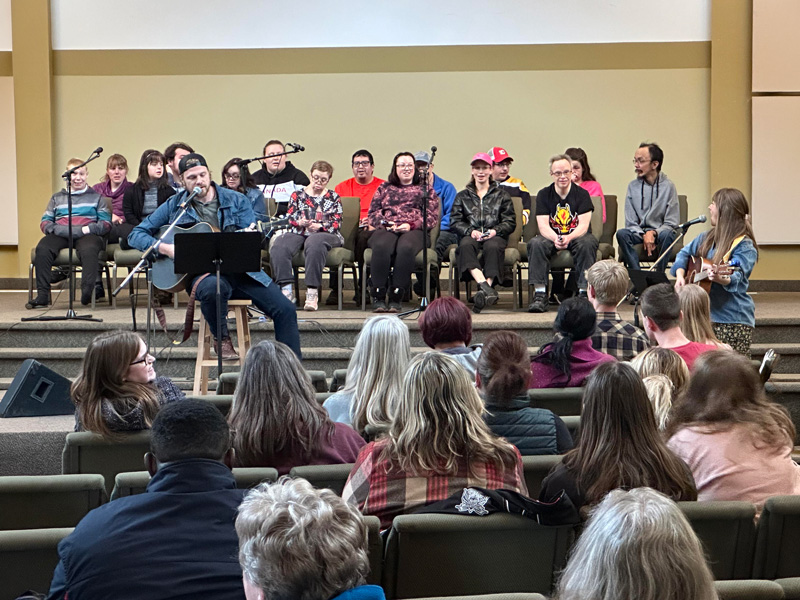 Community-Concert-Music-Series-2 A HopeBridges-style community music gathering in a hall: musicians lead with guitar and microphone while a group of participants sit on stage, and an audience listens from the pews, highlighting connection, inclusion, and support