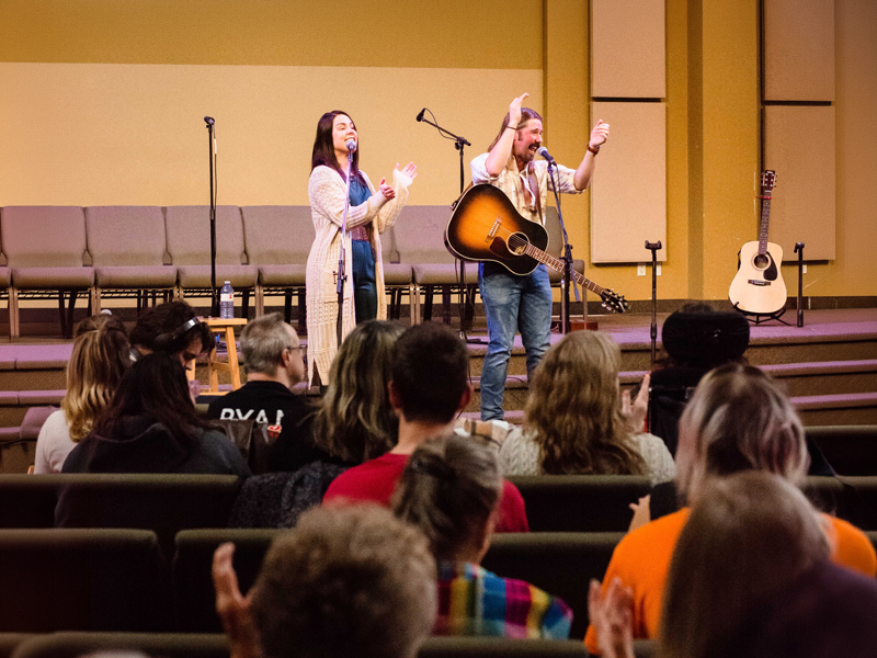 Community-Concert-Music-Series-3 Two music leaders on a small stage with microphones and an acoustic guitar invite audience participation as a seated crowd claps along, creating a warm, community-focused gathering.