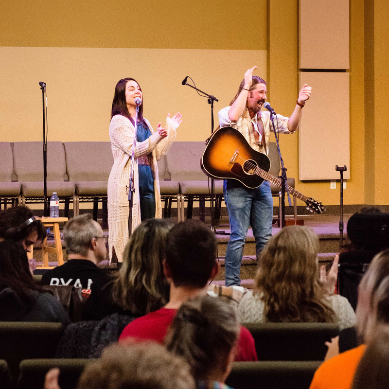 Two music leaders on a small stage with microphones and an acoustic guitar invite audience participation as a seated crowd claps along, creating a warm, community-focused gathering.