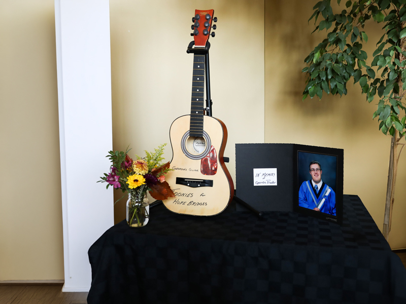 Hope Bridges – Artists Among us Gala Memorial fundraising display with a guitar on a stand labelled “Donations for Hope Bridges,” a framed tribute photo and note “In memory of Gordon Franks,” and a vase of flowers.