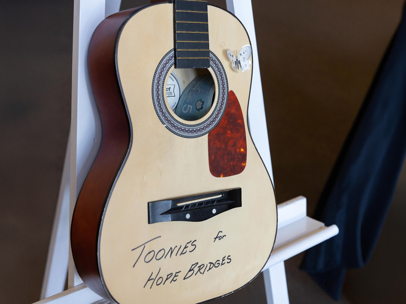 Donate_3 Acoustic guitar on a white stand with cash in the sound hole and handwritten text reading “Toonies for Hope Bridges,” indicating a donation fundraiser.