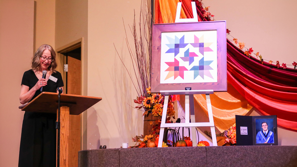 Hope Bridges – Artists Among us Gala Two people stand on a stage beside an easel displaying a framed colourful geometric artwork during HopeBridges (hopebridges.ca) past honours recognizing Christene Howard.