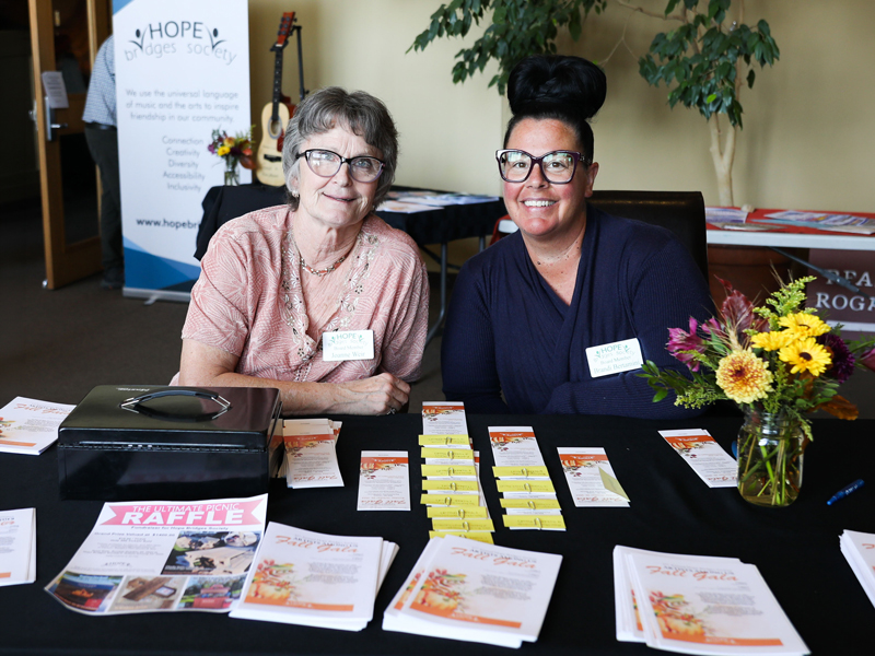 Hope Bridges – Artists Among us Gala Two people at a Hope Bridges Society outreach table with brochures, name tags, a raffle display, and flowers; a guitar and Hope Bridges banner are visible behind them.