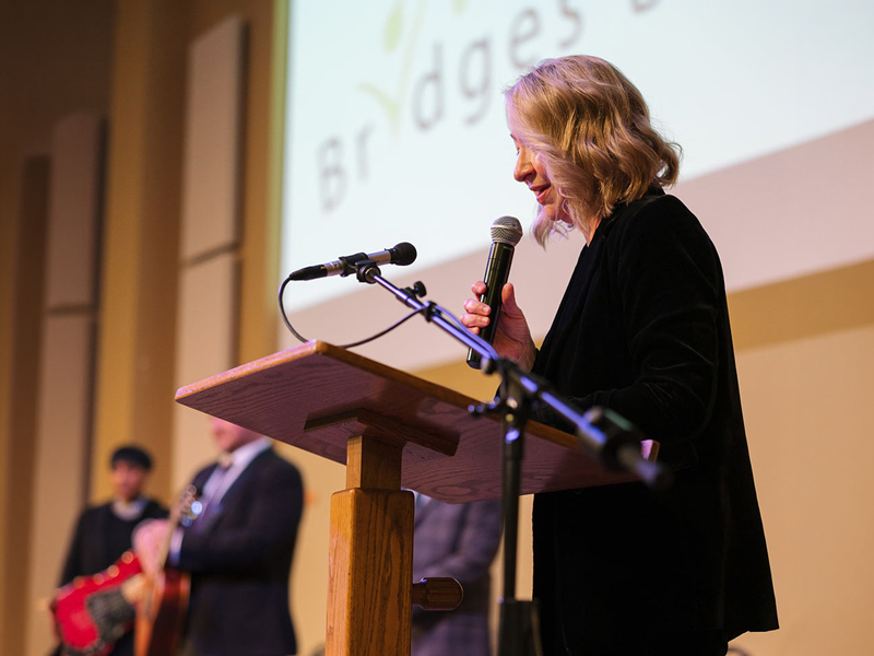 Hope Bridges – Artists Among us Gala Speaker at a podium holding a microphone during a Hope Bridges event, with musicians blurred in the background.