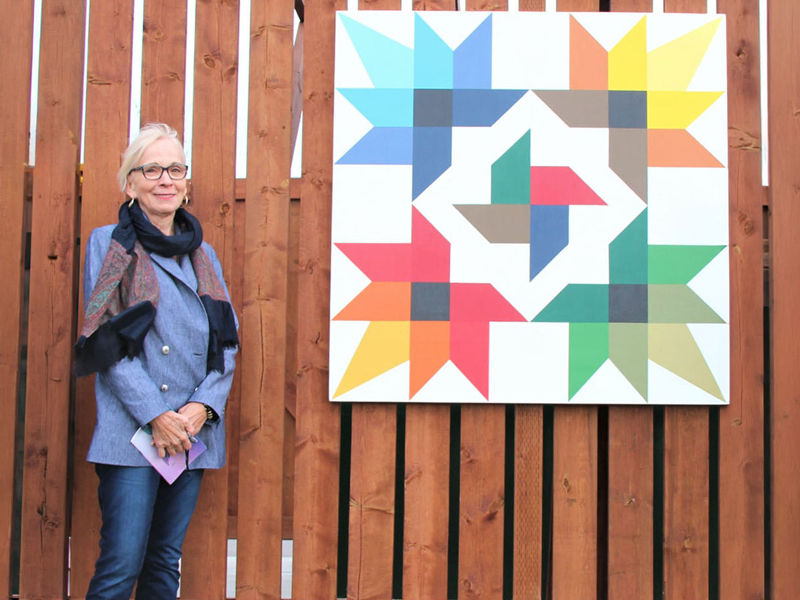 Join-The-Board-Of-Directors-3 Person beside a colourful quilt-pattern mural on a wooden fence, representing community connection.