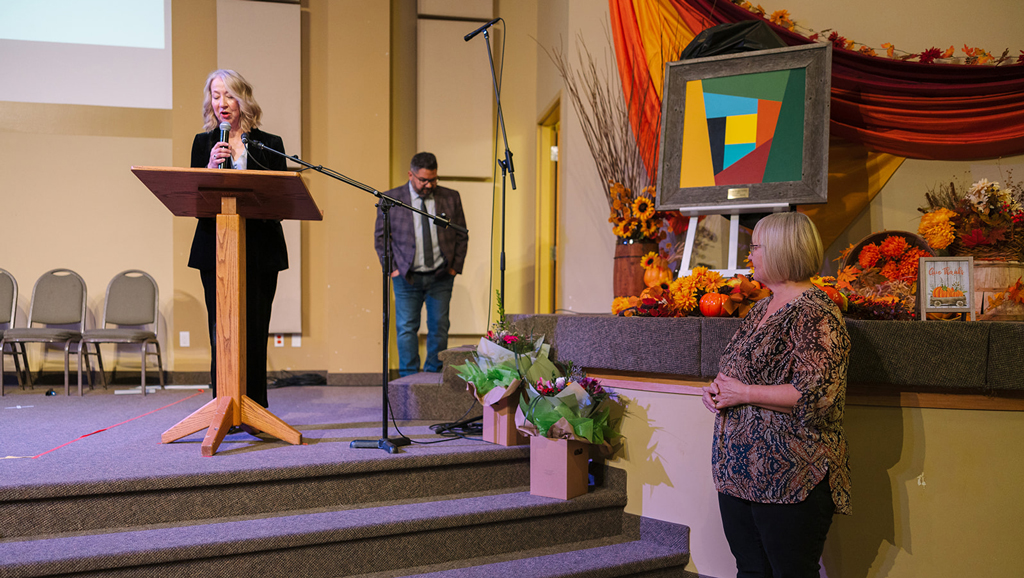 Hope Bridges – Artists Among us Gala Speaker at a podium during HopeBridges (hopebridges.ca) Past Honours recognizing Linda Ray, with an abstract framed artwork on an easel, fall décor, and flower arrangements on the stage.