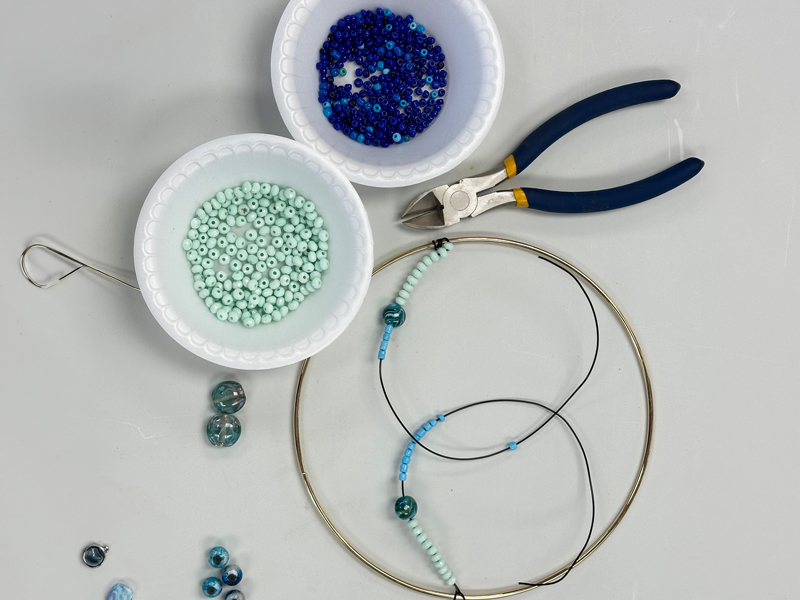 Make-&-Mingle_1 Beading supplies for a “Make & Mingle” craft: bowls of blue and mint seed beads, pliers and wire cutters, and a partly strung bracelet on wire and cord laid out on a tabletop.