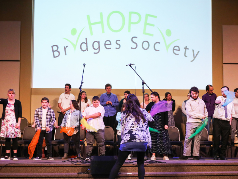 Moving-&-Grooving_3 A “moving and grooving” group stands on a stage holding colourful scarves while a facilitator leads from the front, with the Hope Bridges Society logo projected behind them.