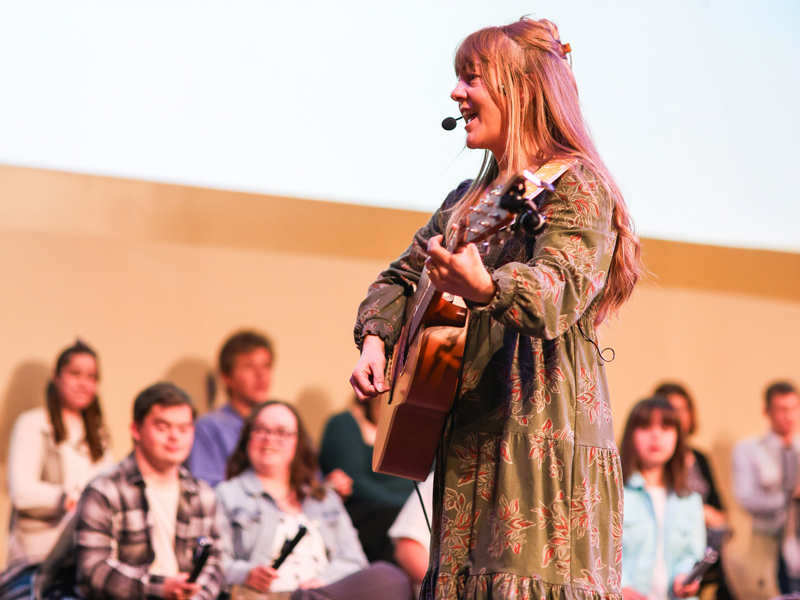 Hope Bridges – Artists Among us Gala A community musician plays an acoustic guitar on stage while people sit and listen behind her, creating a welcoming, HopeBridges-style space for connection and encouragement.
