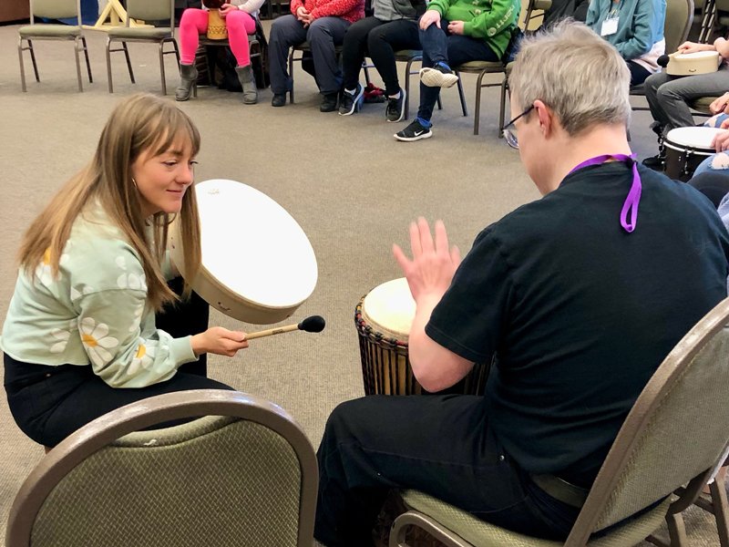 Music-Discovery_2 A facilitator and a participant sit facing each other in a HopeBridges-style music circle, playing hand drums together while others watch from chairs around the room.