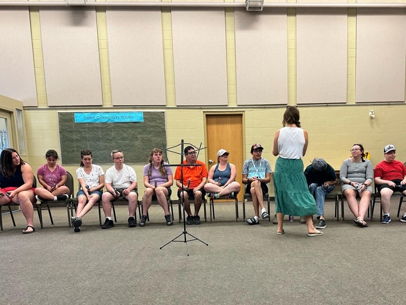 Music-Discovery_3 A HopeBridges-style youth group session in a community hall: a facilitator stands facing a row of seated young people, with a music stand in the centre, creating a calm, supportive space for connection.
