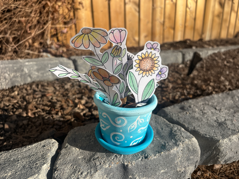 Paper-Flowers-in-Pot Small turquoise flowerpot with white swirl patterns holding hand-drawn paper flowers, sitting on a stone in a garden bed in front of a wooden fence.