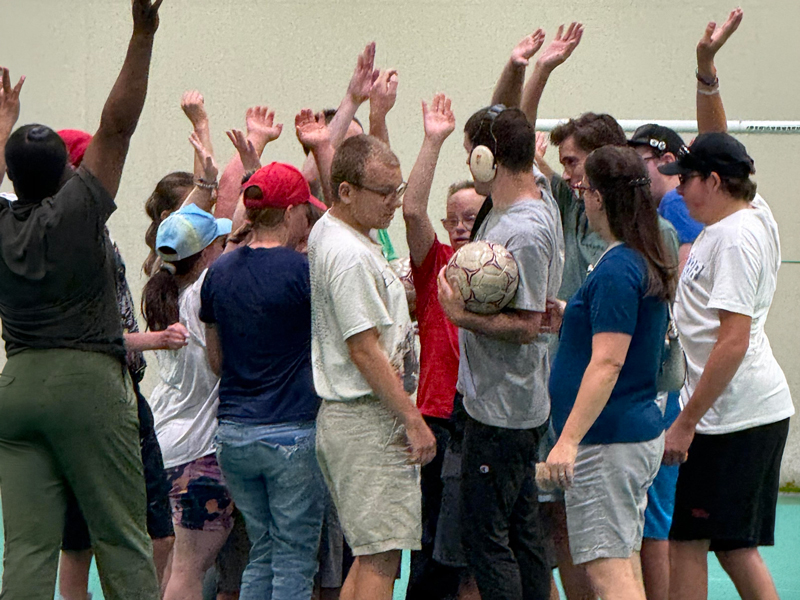 Way2Move_1 Adults in an adaptive sports group gather in a gym with hands raised during a team huddle; one participant holds a soccer ball, reflecting HopeBridges Way2 Move inclusive recreation.